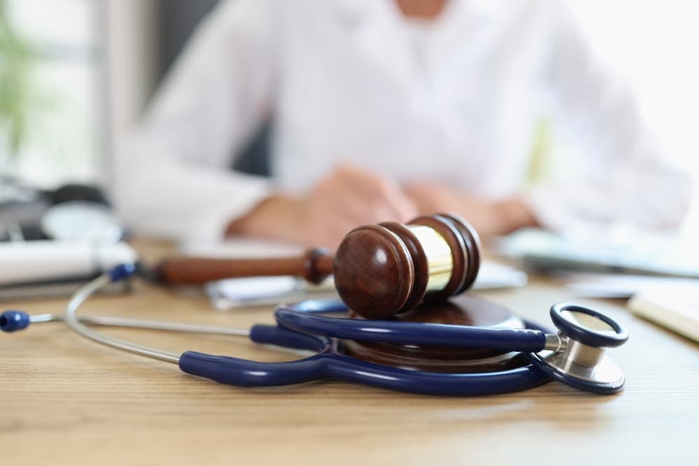 Closeup of judge's gavel with a stethoscope and doctor writing notes in the background. Represents the legal aspects of healthcare, medical insurance, and healthcare disputes.