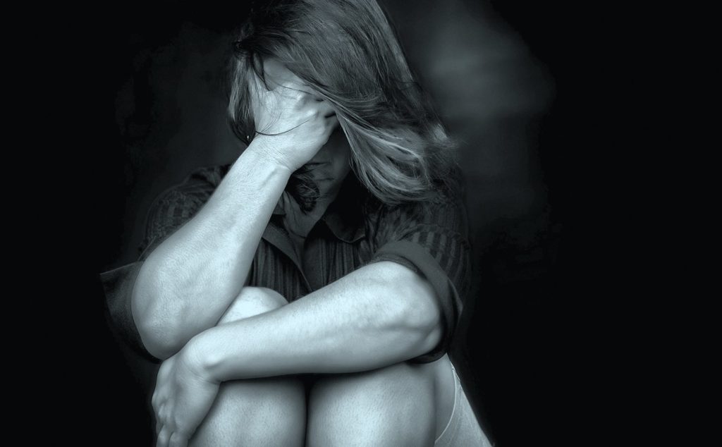 A distressed person sitting with knees drawn up, covering their face with one hand, symbolizing sadness or trauma.