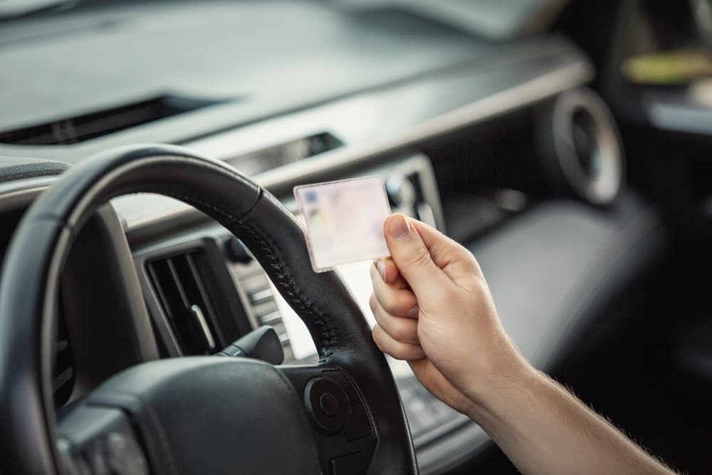 A close-up shot of a person's hand holding a driver's license in front of a steering wheel.