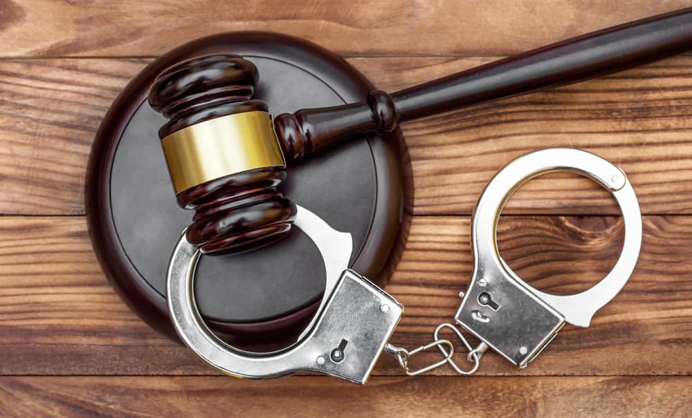 Top view of a wooden courtroom desk with a gavel, stand, and handcuffs placed together, symbolizing legal proceedings and justice.