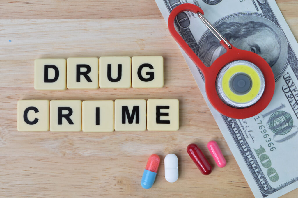 The Importance of Legal Representation in Drug Cases Wooden surface with the words 'DRUG CRIME' spelled out in letter tiles, U.S. hundred-dollar bill partially visible, a red keychain flashlight, and several colorful pills arranged at the bottom.