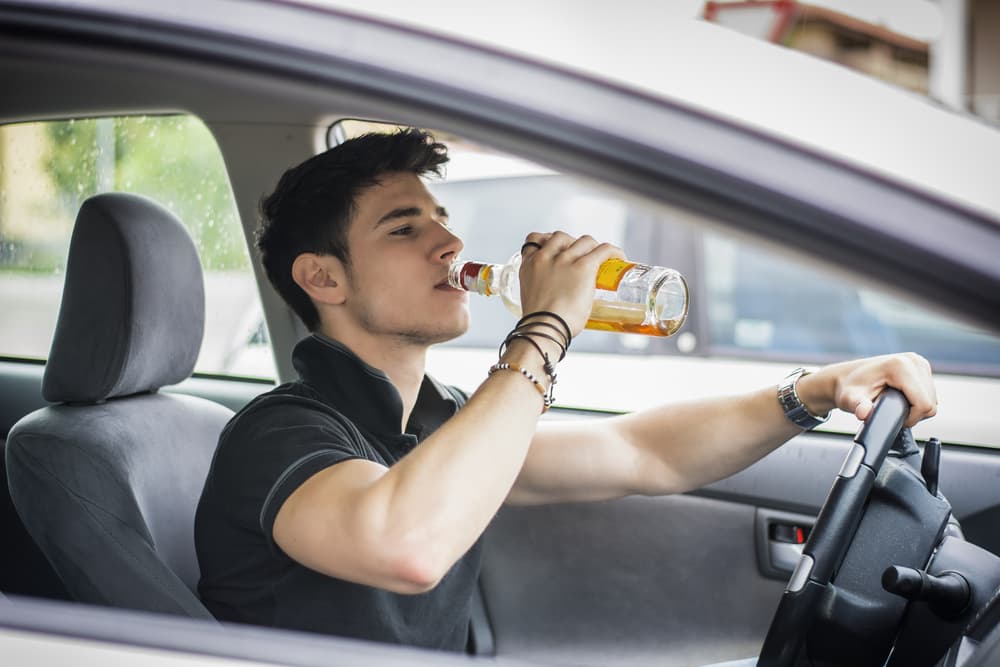 A description of a risky scenario depicting a young man drinking alcohol while driving in traffic.
