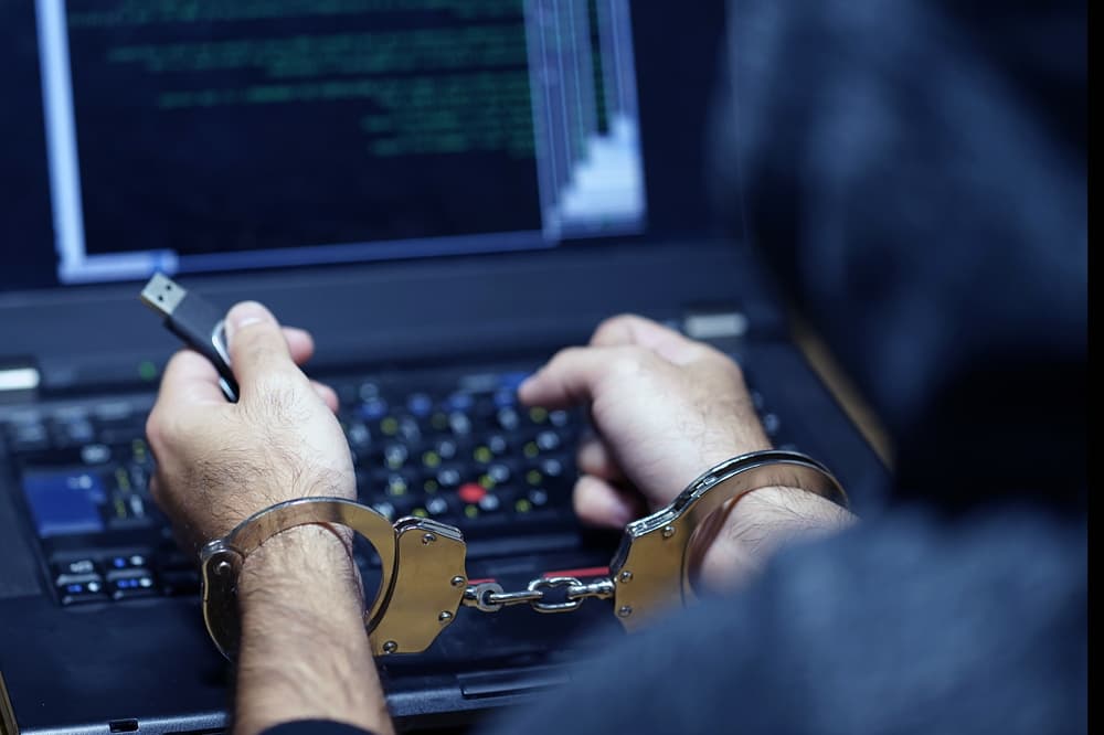 Back view of a hacker in handcuffs holding a USB drive, with a selective focus on their hands. Depicts a prisoner or arrested individual.