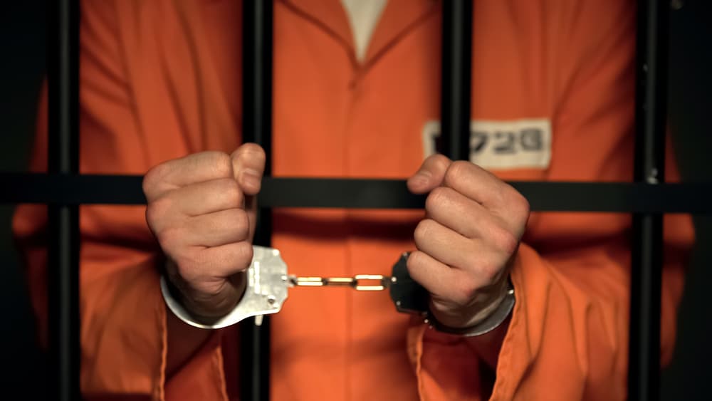 A prisoner displaying their hands in handcuffs, a menacing criminal positioned behind bars.






