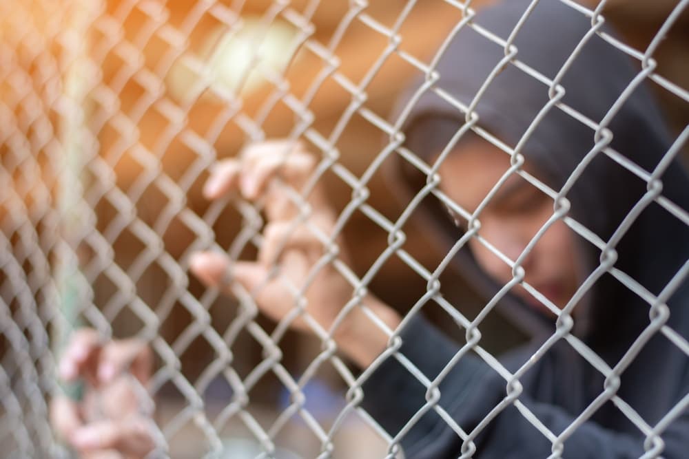 A young boy in a gray hoodie stands with his hands on a metal fence in a detention center, looking sorrowful and longing for freedom.