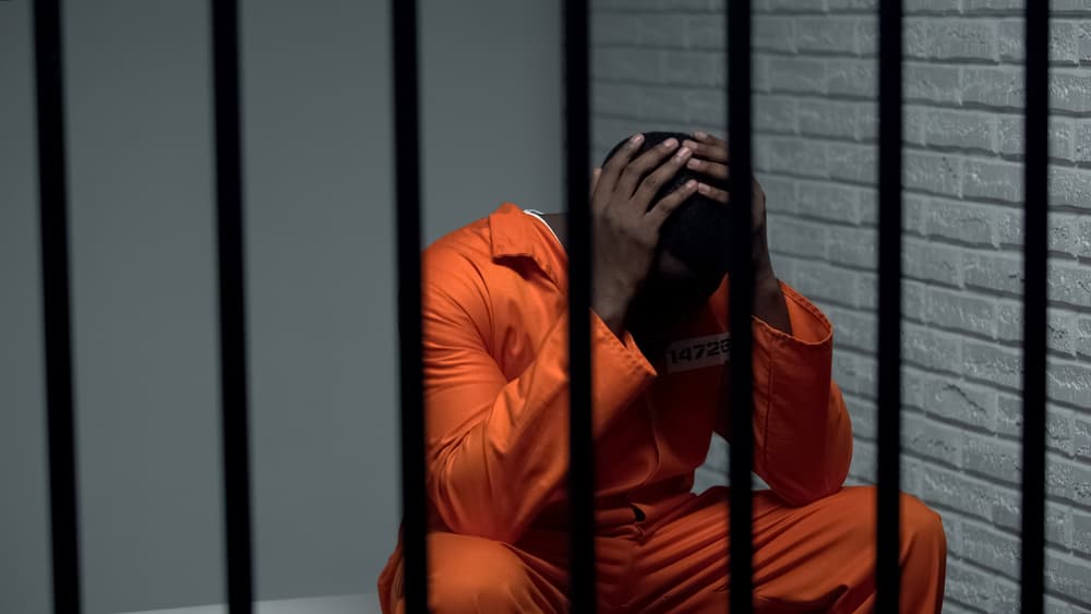 A distressed Black prisoner sits in a cell, reflecting the plight of a wrongly accused individual and the flaws within the justice system.