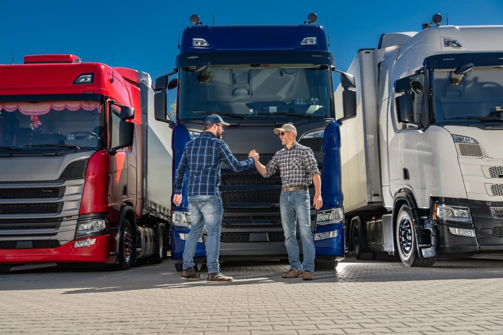 Two Caucasian semi-truck drivers shake hands in front of a lineup of Euro trucks, symbolizing partnership and teamwork in the transportation industry.







