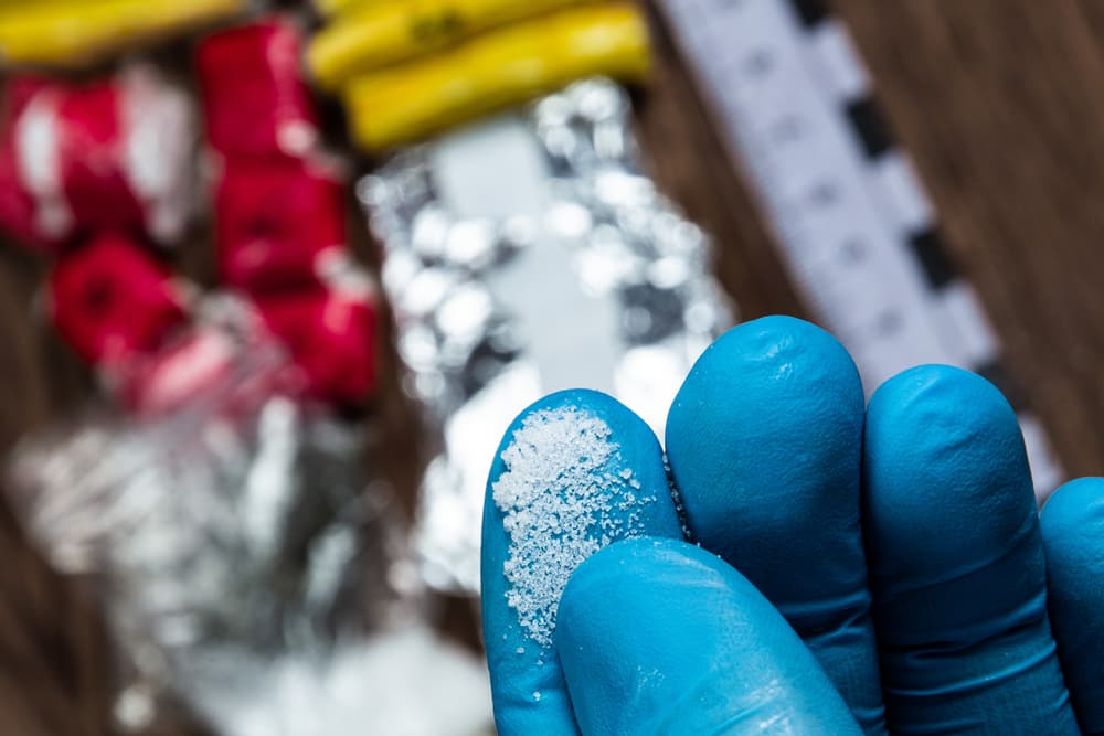 Gloved hand holding white powder, with drug packages and measuring scale behind.